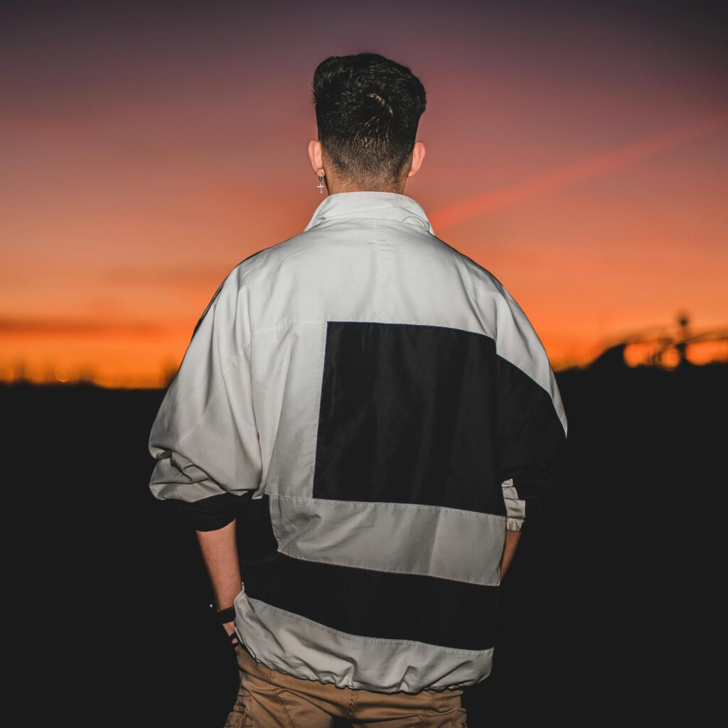 A man in an oversized jacket standing outdoors during twilight, with a vibrant sunset in the background.